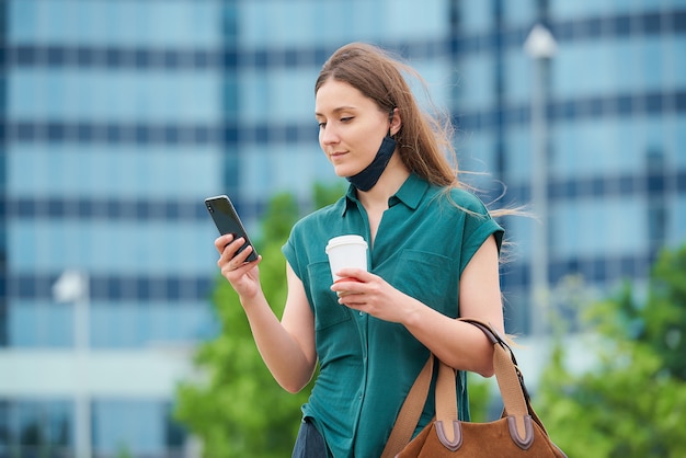 A young woman in a deflated medical face mask is reading the news on a smartphone and drinking coffee in downtown