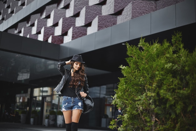 Photo young woman in denim shorts, black hat and leather jacket with black leather backpack walking in the street of european city. trendy casual outfit. everyday look. street fashion.