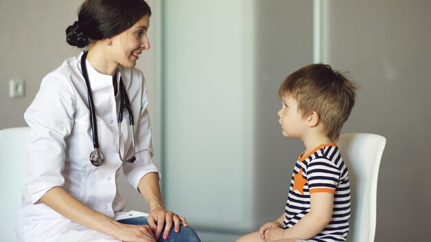 Young woman doctor talking little boy and listening with stethoscope in medical office