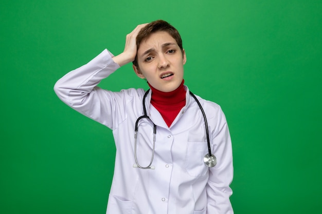 Young woman doctor in white coat with stethoscope looking confused holding hand on her head for mistake