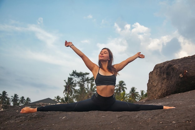 Young woman does yoga position splits or twine for healthy lifestyle on tropical coast sea looking away arms raised