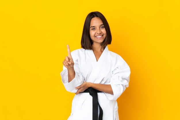 Young woman doing karate isolated on yellow wall showing and lifting a finger