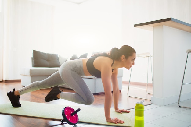 Young woman doing sport workout in room during quarantine. Exercising in room on yoga mat. Stand in plank position and and doing mount climber exercise.