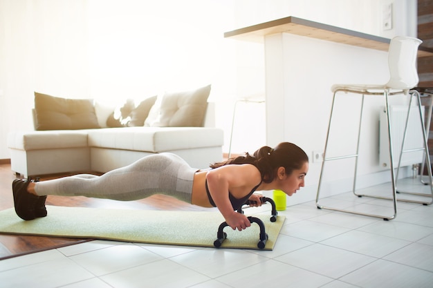 Young woman doing sport workout in room during quarantine. Fitness model stand in plank position using push up stand hand bar. Also pushing up on mat.