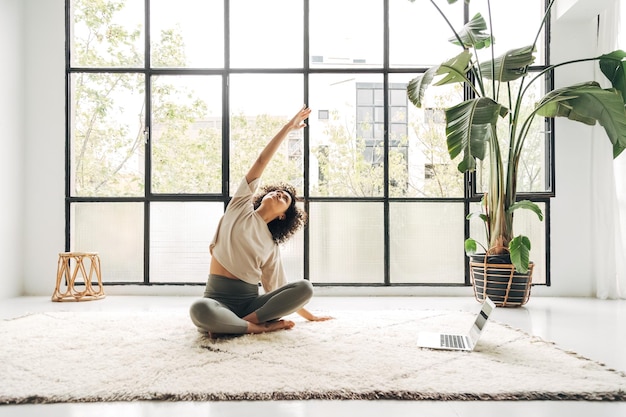 Young woman doing stretching exercises at home following online fitness video with laptop Copy space Wellness and healthy lifestyle concept