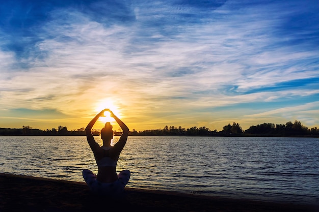 Young woman doing yoga exercises on the lake beach at sunset