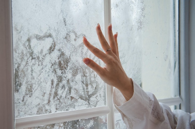 Young woman drawing heart on a foggy window. A beautiful female hand draws a heart on a misty window.