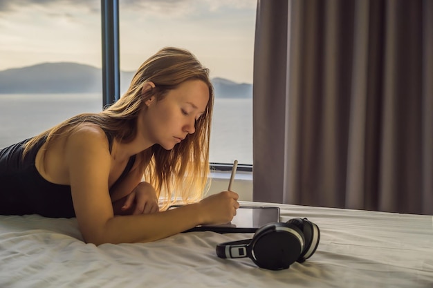 Young woman draws on a tablet against the background of a window with a sea view Designer sketching