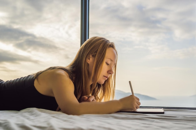 Young woman draws on a tablet against the background of a window with a sea view Designer sketching