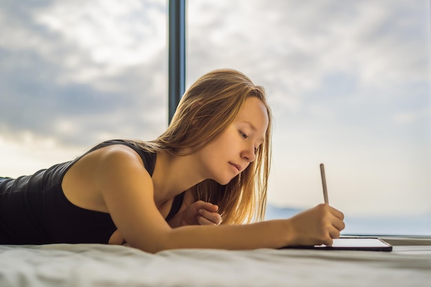 Young woman draws on a tablet against the background of a window with a sea view Designer sketching