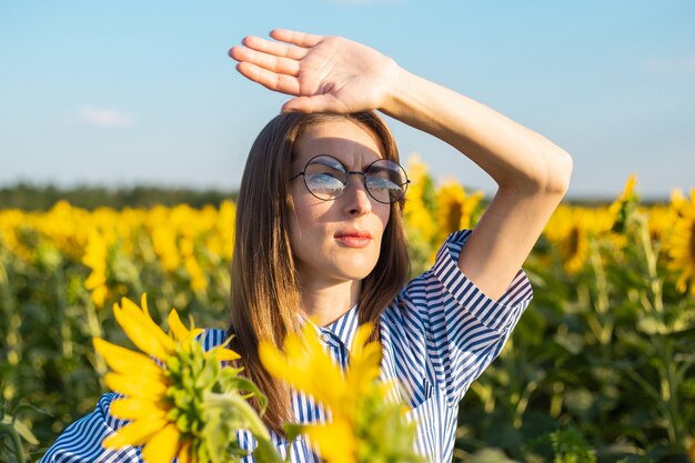 Young woman in dress and glasses greets the sunrise and covers her face on a sunflower field.