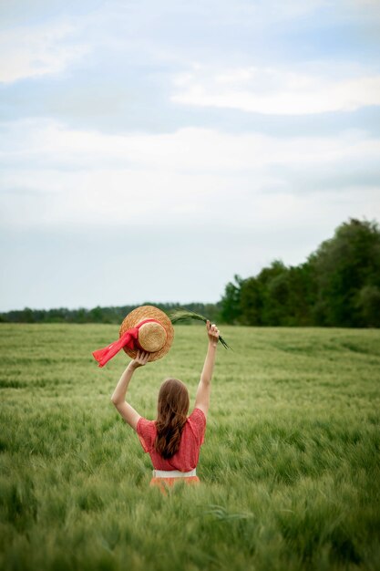 Young woman in dress and hat in green field of barley in countryside. Stylish girl in rustic dress enjoying peaceful moment in grass in summer. Tranquil rural moment