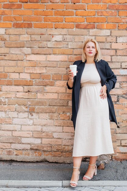 Young woman in a dress and jacket stands by a brick wall and drinks coffee