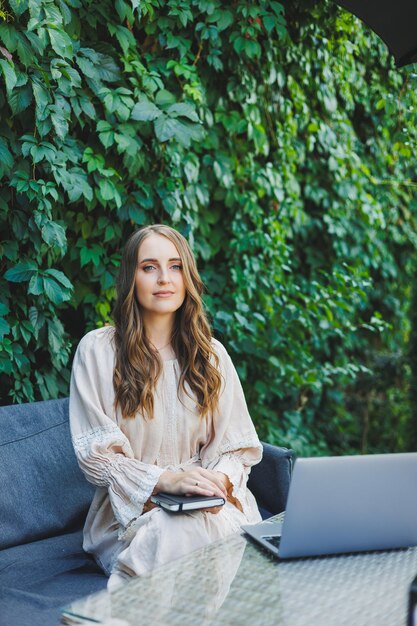 A young woman in a dress sits with a laptop and makes notes on the terrace of a summer cafe Woman in casual stylish summer clothes