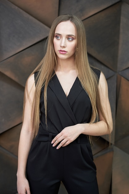 Young woman dressed in black jumpsuit, heeled shoes posing . Beautiful blonde girl demonstrating stylish smart clothing against wall