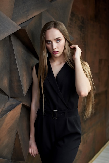 Young woman dressed in black jumpsuit, heeled shoes posing . Beautiful blonde girl demonstrating stylish smart clothing against wall