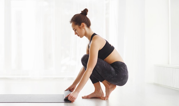 A young woman dressed in black sportswear performs yoga rolling up fitness mat the bright airy room