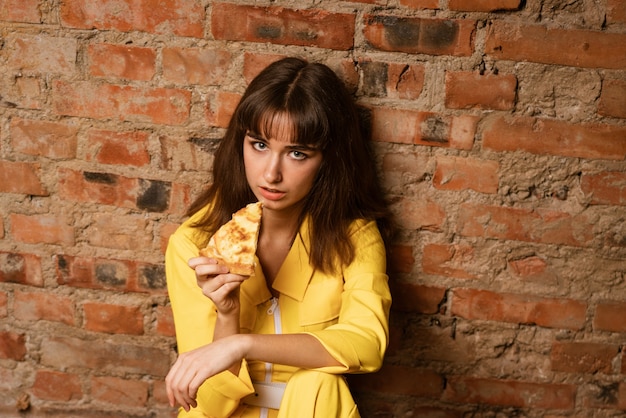 A young woman dressed in a yellow suit eats a slice of pizza leaning against a brick wall.