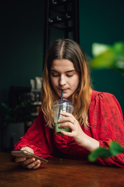 Young woman drinking green drink ice matcha latte in cafe and using smartphone