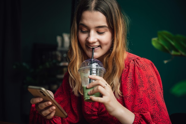 Young woman drinking green drink ice matcha latte in cafe and using smartphone