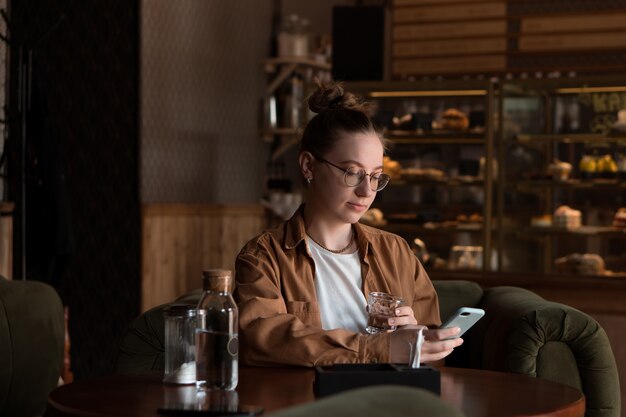 Young woman drinking water in cafe waits for ordered food using mobile phone