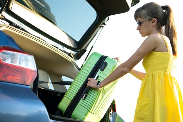 Young woman driver taking green suitcase out from car trunk. Travel and vacations concept.