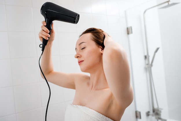 Young woman drying and styling hair with hair dryer making hairstyle in modern bathroom interior Beauty routine after morning shower