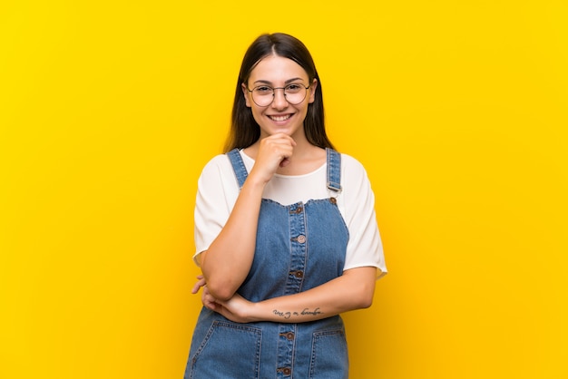 Young woman in dungarees isolated on yellow  with glasses and smiling