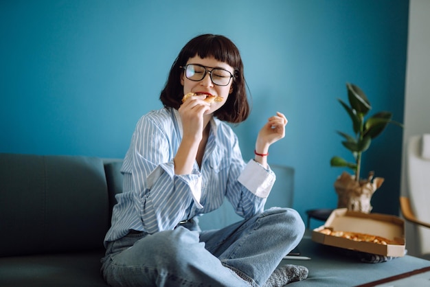 Young woman eating a piece of delicious pizza from the box sitting at sofa at home Food delivery