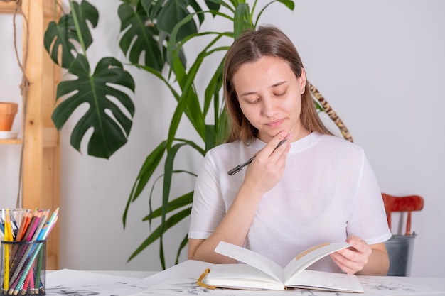 A young woman of European appearance does homework at school university The girl diligently studies at home at a white table Photography in light shades in a home interior