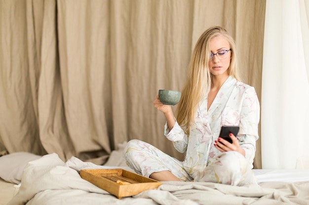 Young woman in eyeglasses checking news in her smartphone in bed during breakfast