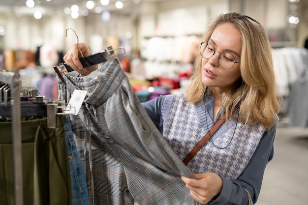 Young woman in eyeglasses holding looking at new trousers in her hands while buying clothes in the store