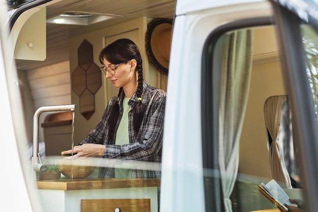 Young woman in eyeglasses washing vegetables under water in sink while standing in the van during ca