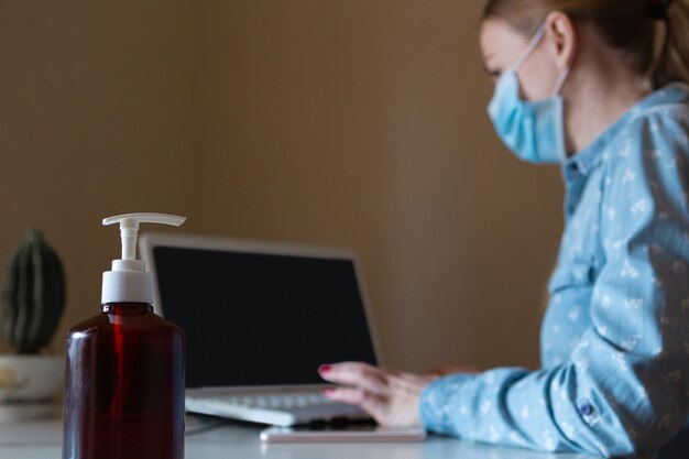 Young woman in face mask disinfecting gadgets surfaces on her workplace