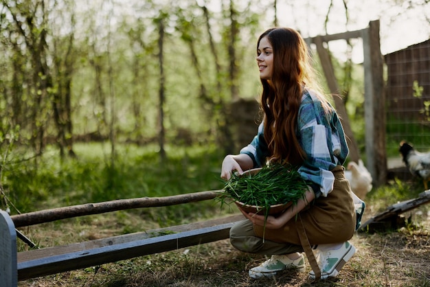 A young woman farmer sits at a bird feeder and checks the composition of the grain to feed the chickens in a pen in the countryside