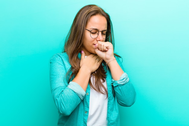 Young woman feeling ill with a sore throat and flu symptoms, coughing with mouth covered over blue wall