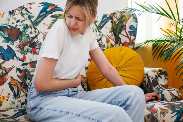 Young woman feels sick in stomach. Unhealthy female, sitting on couch and holding her stomach, feeling uncomfortable. Concept of health problems, food poisoning, gastritis. Selective focus on hands