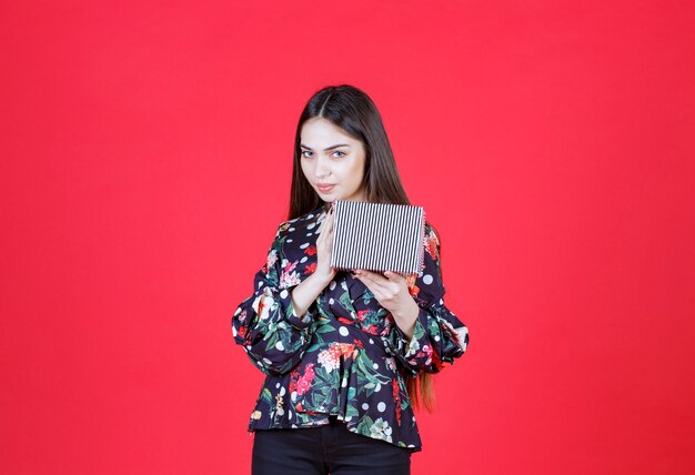 Young woman in floral shirt holding a silver gift box and looks thoughtful