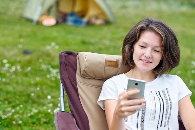 Young woman freelancer sitting on chair and using smart phone Relaxing in forest or meadow Remote work and outdoor activity in summer