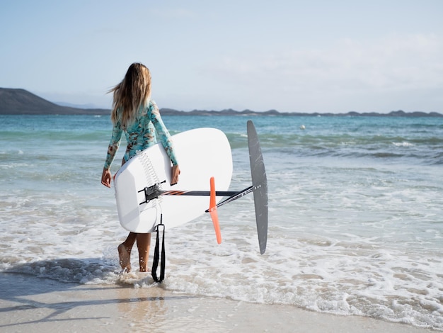 Young woman from behind walking to the water on the beach shore to practice hydrofoil surfing water