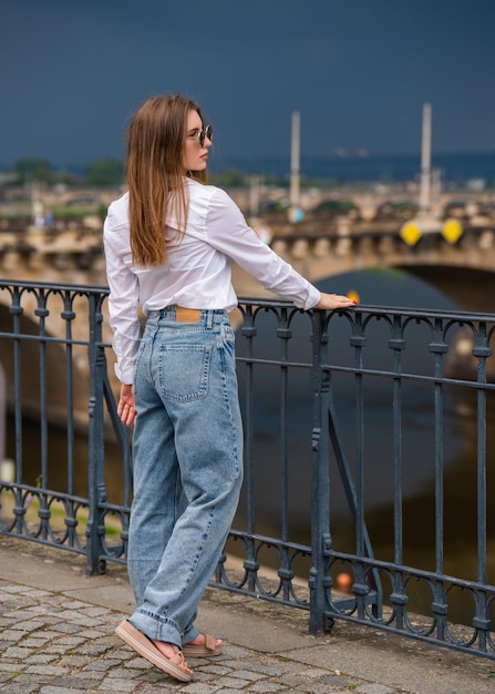 Young woman in full growth The girl stands against the backdrop of the river holding the railing and leaning back Look up Back view