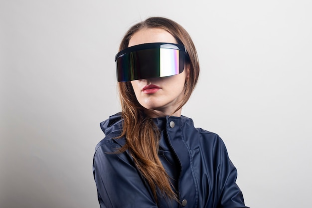 Young woman of the future in virtual reality glasses and in a blue jacket on a light background.