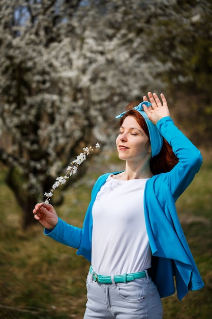 A young woman girl stands in a garden where cherries and apricots blossom. She is happy breathing air, spring flowers in the background