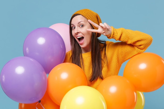 Young woman girl in sweater hat posing isolated on blue background. Birthday holiday party, people emotions concept. Mock up copy space. Celebrating hold colorful air balloons showing victory sign.