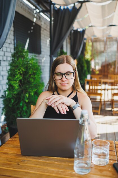 A young woman in glasses and a black dress sits on the summer terrace of a cafe and works at a laptop Female freelancer works remotely online while sitting in a summer cafe