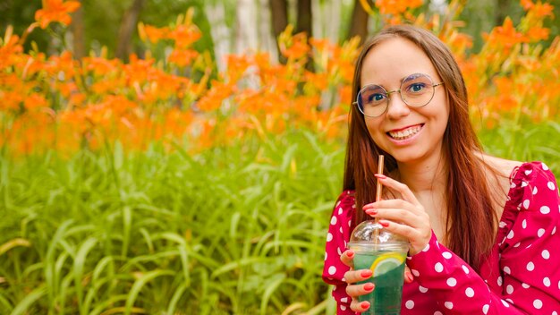 Young woman in glasses drinking refreshing lemonade sitting in city park Relaxed female in red dress drinking soft drink through straw quenching thirst on background of blurred flowers