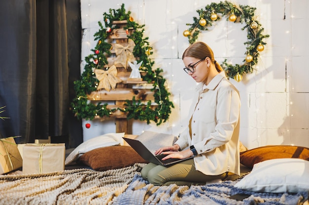 A young woman in glasses sits on a bed in a home environment and works remotely New Year New Year's decor in the room