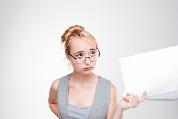 Young woman in glasses with papers in hand feels disappointed, grieved, sad and chagrined. Failure, bad luck, life or business problems. Grey wall with copy space