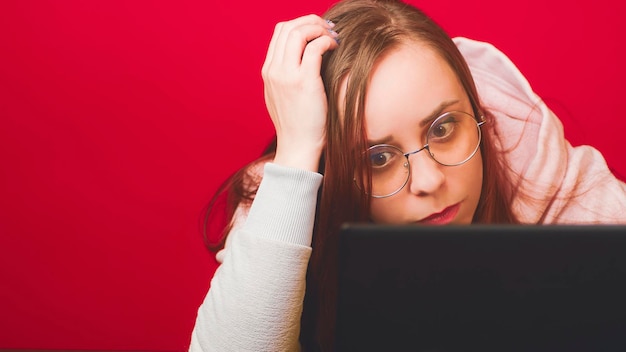 Young woman in glasses works on laptop and scratches head sitting in home office Business freelancer works remotely at modern workplace on red background