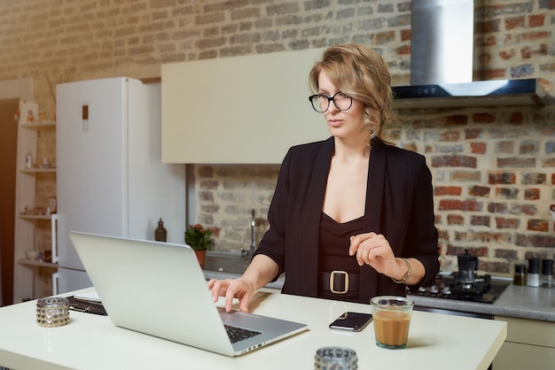 A young woman in glasses works remotely on a laptop in her kitchen. A serious girl calmly browsing news on the internet at home.